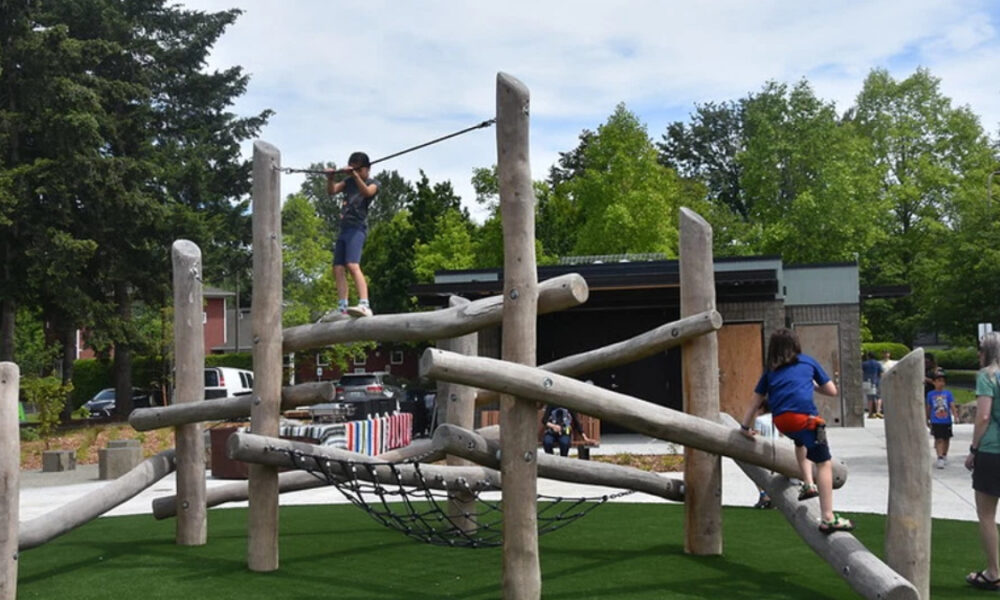 Children climb on a wooden jungle gym outdoors on green turf with trees in the background.