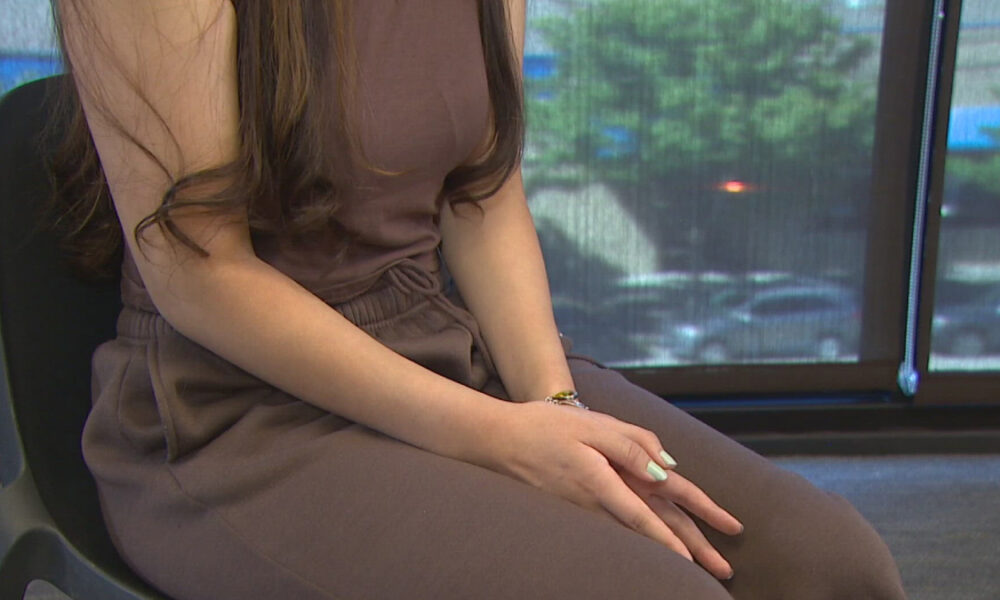 Person seated in a brown outfit with hands gently folded on their lap, by a window with a blurred outdoor view.