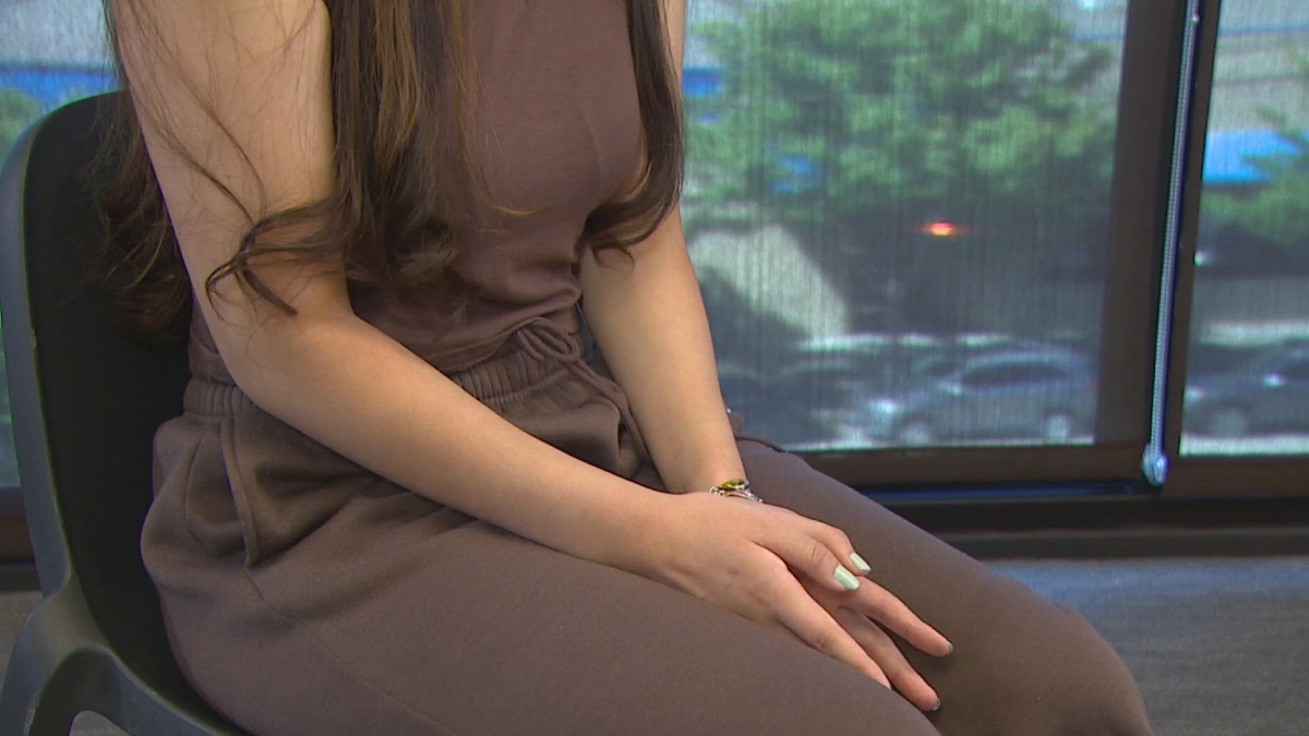 Person seated in a brown outfit with hands gently folded on their lap, by a window with a blurred outdoor view.