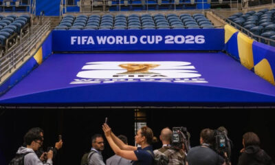 Cameras and reporters photograph a blue FIFA World Cup 2026 entrance canopy with the event logo.