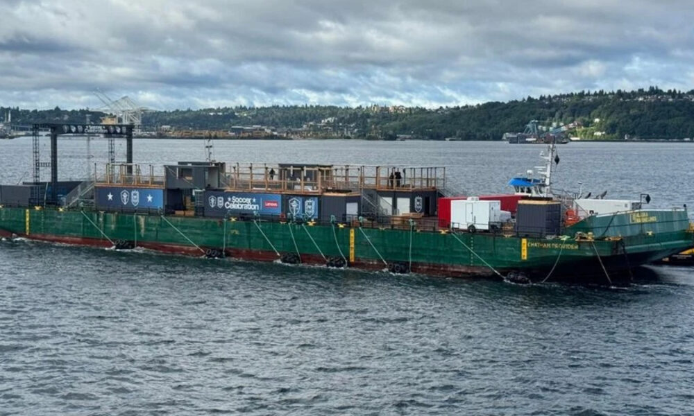 Green event barge on a body of water with a wooden deck and banners for a 'Soccer Celebration' event, anchored with ropes; a distant shoreline in the background.