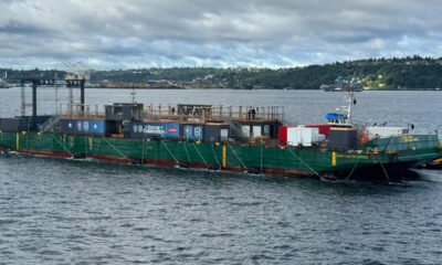 Green event barge on a body of water with a wooden deck and banners for a 'Soccer Celebration' event, anchored with ropes; a distant shoreline in the background.