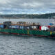 Green event barge on a body of water with a wooden deck and banners for a 'Soccer Celebration' event, anchored with ropes; a distant shoreline in the background.