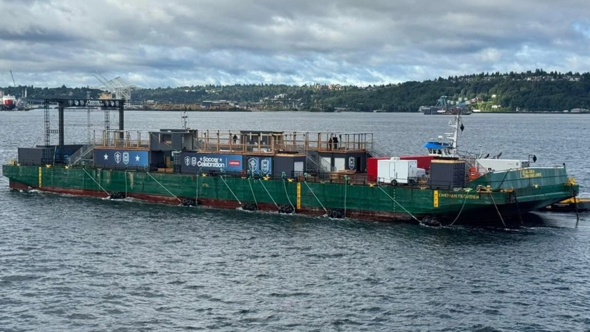 Green event barge on a body of water with a wooden deck and banners for a 'Soccer Celebration' event, anchored with ropes; a distant shoreline in the background.