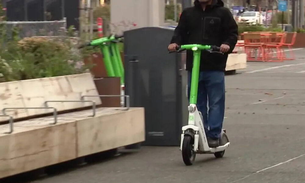 Person in a dark jacket rides a bright green and white electric scooter along a city street with planters nearby