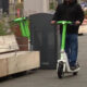 Person in a dark jacket rides a bright green and white electric scooter along a city street with planters nearby