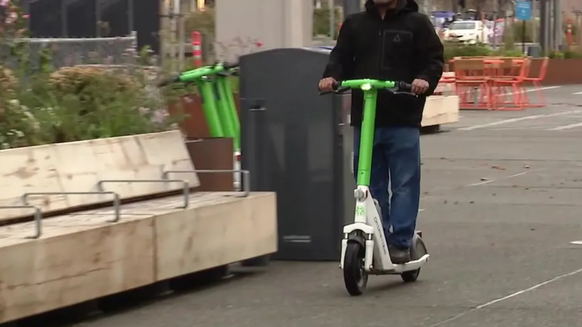 Person in a dark jacket rides a bright green and white electric scooter along a city street with planters nearby