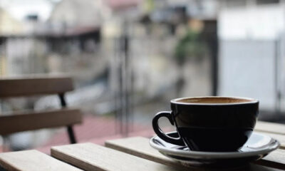 Black coffee cup on a saucer resting on a wooden outdoor table with a blurred urban background while seated at a cafe.