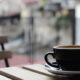 Black coffee cup on a saucer resting on a wooden outdoor table with a blurred urban background while seated at a cafe.