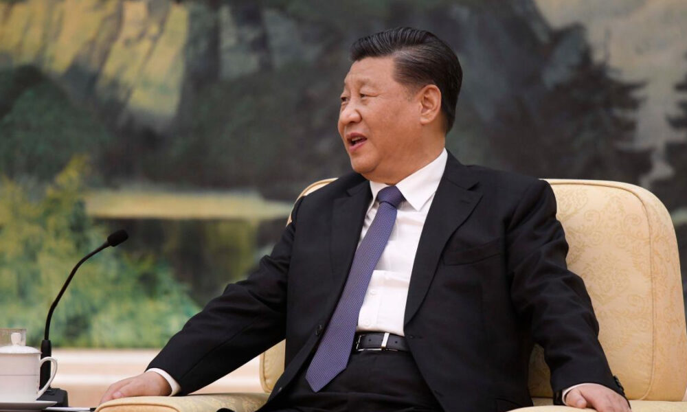 Xi Jinping sits in a cream armchair, wearing a black suit and purple tie, speaking into a microphone.