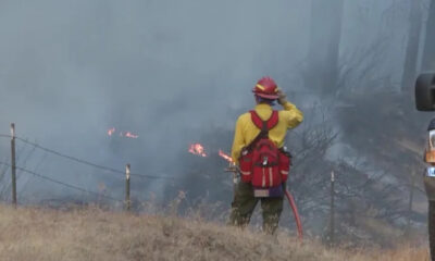 Firefighter in yellow uniform and red helmet stands amid smoke, facing a small grass fire in a rural field with a fence nearby.