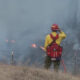 Firefighter in yellow uniform and red helmet stands amid smoke, facing a small grass fire in a rural field with a fence nearby.