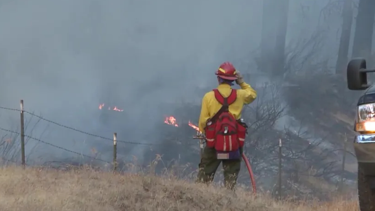 Firefighter in yellow uniform and red helmet stands amid smoke, facing a small grass fire in a rural field with a fence nearby.