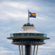 Seattle Space Needle with rainbow Pride flag atop, against a cloudy sky.
