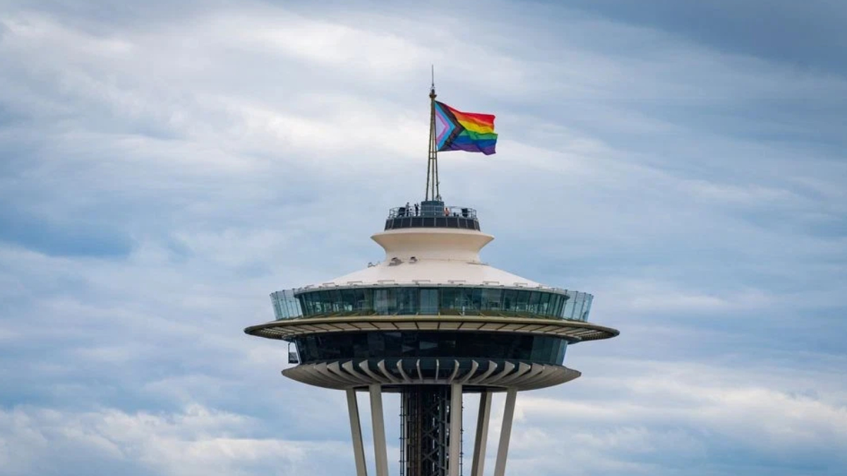 Seattle Space Needle with rainbow Pride flag atop, against a cloudy sky.