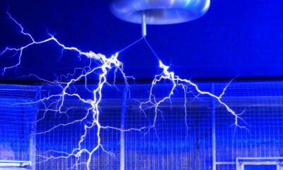 Blue-lit lab with high-voltage electrical arcing across a metal enclosure from a ceiling-mounted device (Tesla coil).