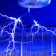 Blue-lit lab with high-voltage electrical arcing across a metal enclosure from a ceiling-mounted device (Tesla coil).