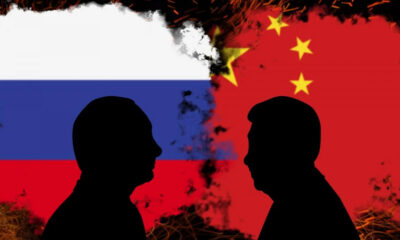 Two black silhouettes face off against a background blending the Russian flag on the left and the Chinese flag on the right, with fiery edges suggesting tension.