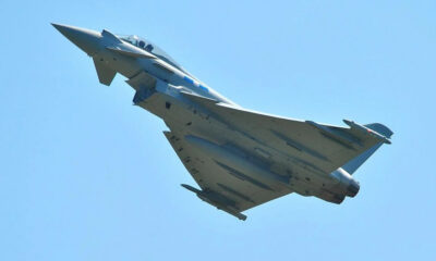 Military fighter jet in flight, viewed from below against a clear blue sky.