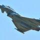 Military fighter jet in flight, viewed from below against a clear blue sky.