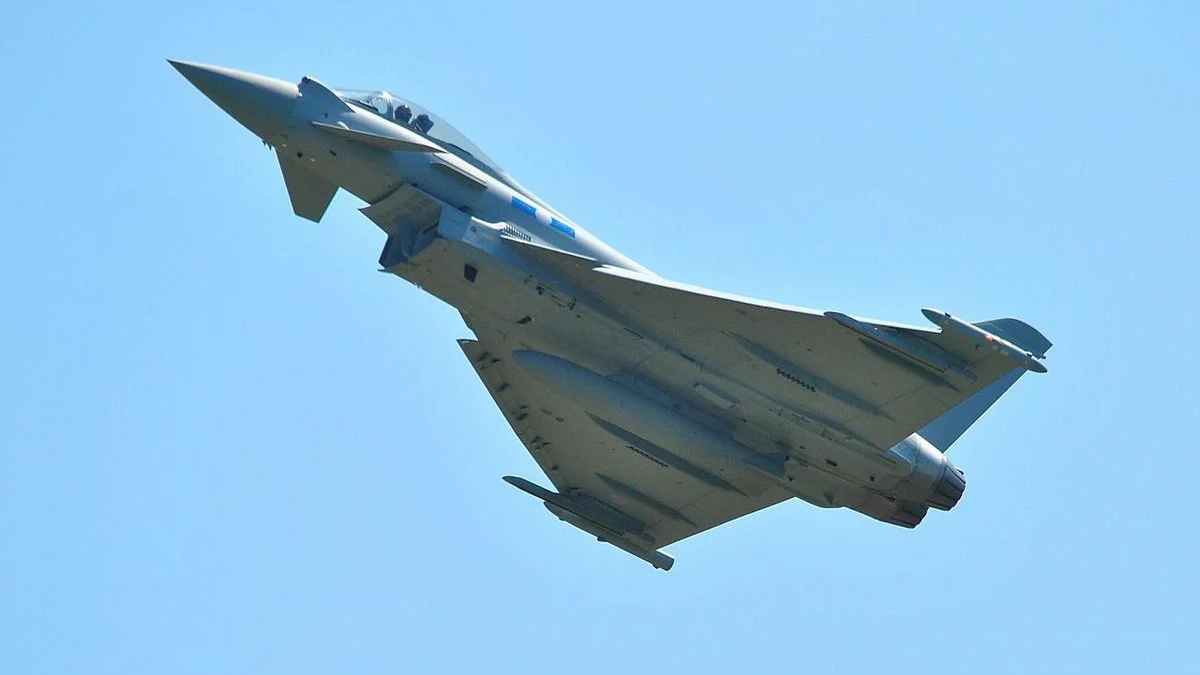 Military fighter jet in flight, viewed from below against a clear blue sky.