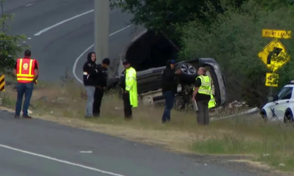 Overturned SUV on a grassy roadside with responders in high-visibility vests and a parked police car nearby.