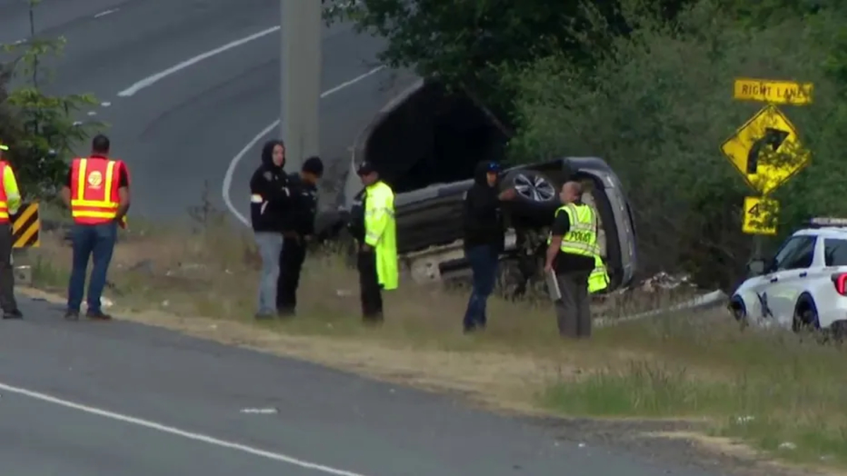 Overturned SUV on a grassy roadside with responders in high-visibility vests and a parked police car nearby.