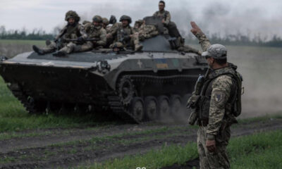 Soldiers ride on a tracked military vehicle in a field as a commander on the ground raises his arm to signal.