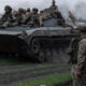 Soldiers ride on a tracked military vehicle in a field as a commander on the ground raises his arm to signal.