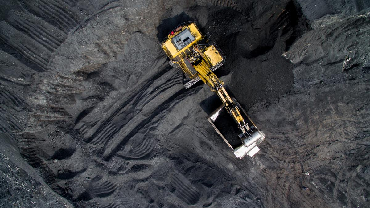 Aerial view of a yellow mining excavator digging a coal pit in dark grey earth, with tire tracks around.