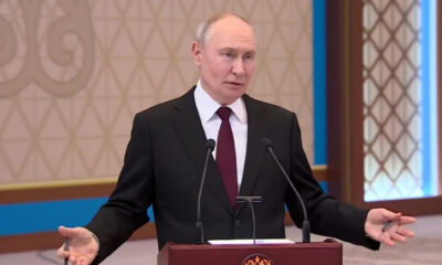 Man in a dark suit and red tie speaks at a wooden podium with two microphones in a formal room.
