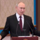 Man in a dark suit and red tie speaks at a wooden podium with two microphones in a formal room.