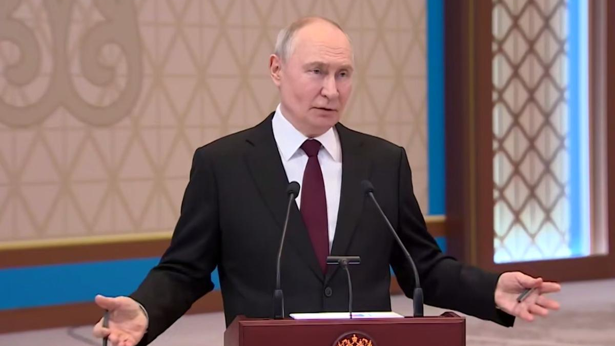 Man in a dark suit and red tie speaks at a wooden podium with two microphones in a formal room.