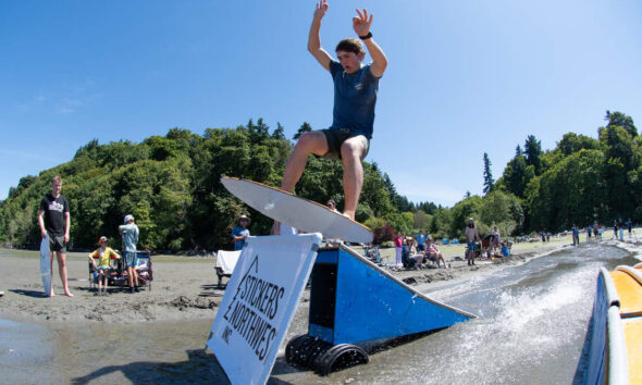 A young man in a blue shirt jumps a wakeboard over a blue ramp into the water while spectators watch on a sunny beach.