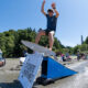 A young man in a blue shirt jumps a wakeboard over a blue ramp into the water while spectators watch on a sunny beach.