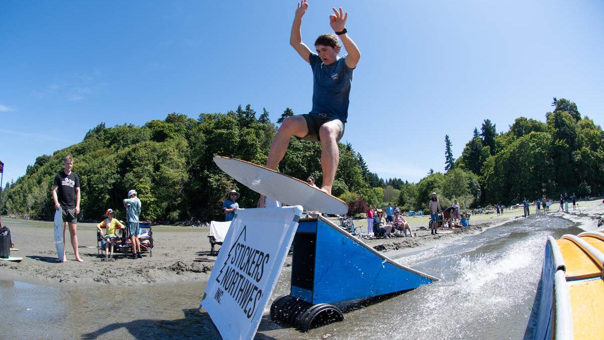 A young man in a blue shirt jumps a wakeboard over a blue ramp into the water while spectators watch on a sunny beach.