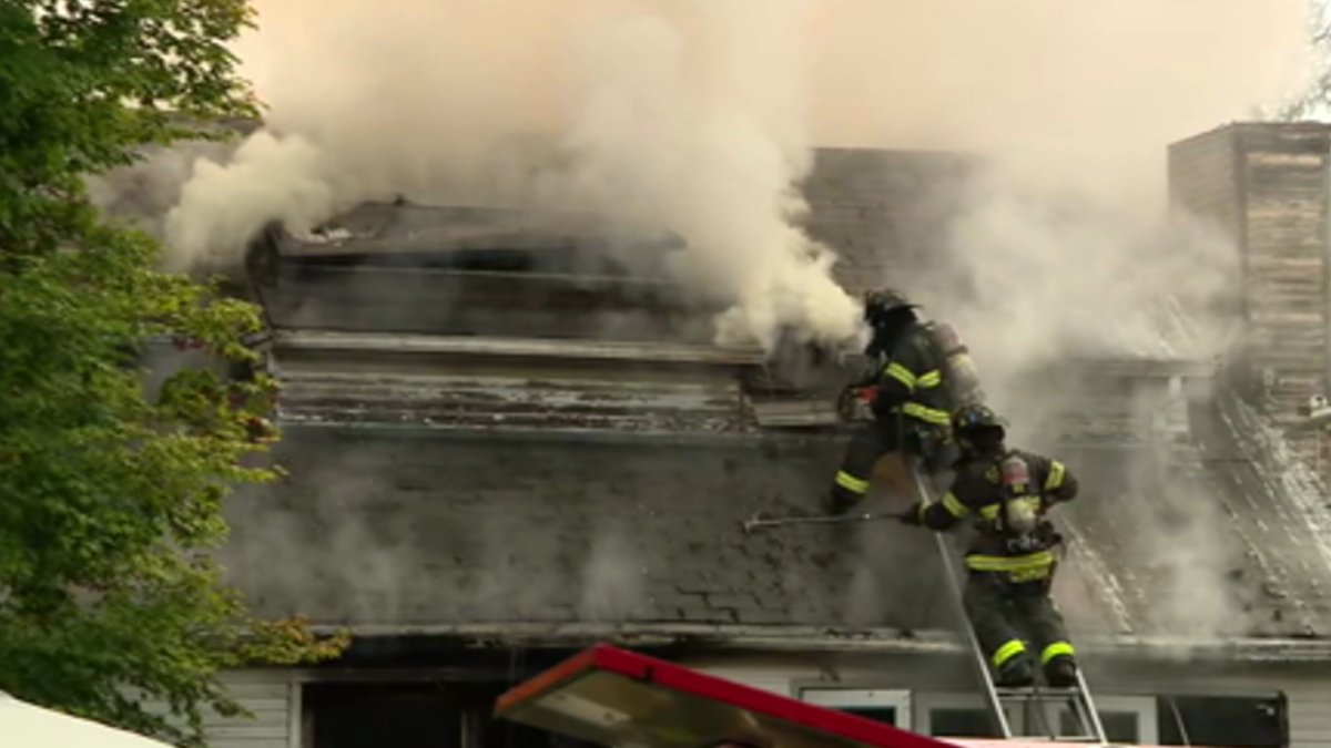 Firefighters on a smoke-filled roof spraying water to extinguish a residential fire.