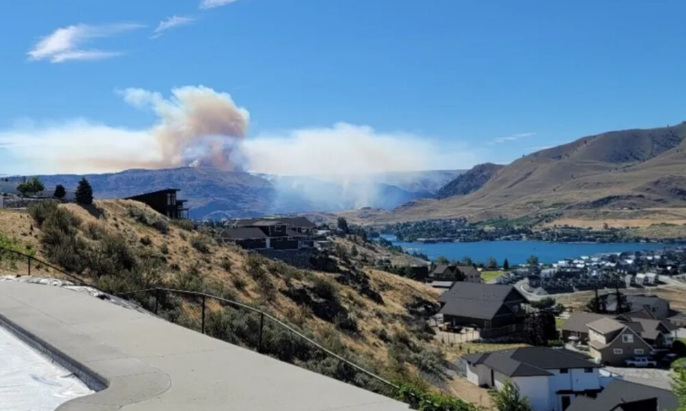 Smoke plume rises from distant hills over a lake town, with houses on a hillside in the foreground.