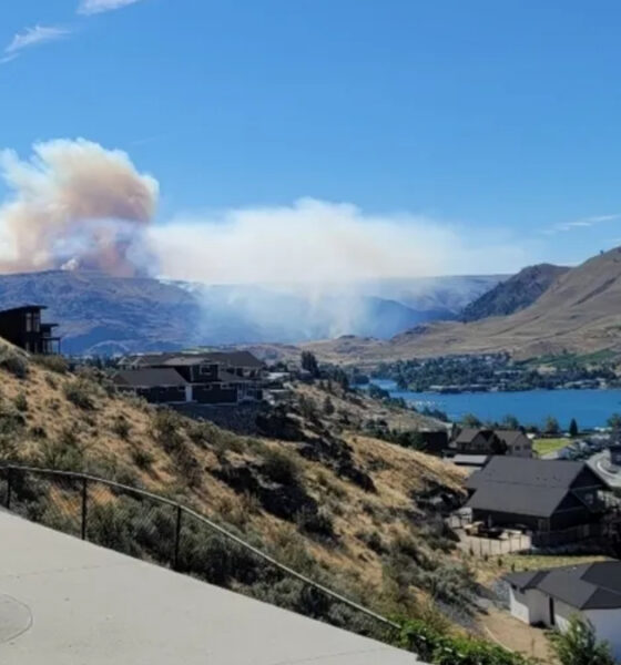 Smoke plume rises from distant hills over a lake town, with houses on a hillside in the foreground.