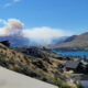 Smoke plume rises from distant hills over a lake town, with houses on a hillside in the foreground.