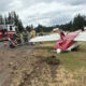 Downed red-and-white small airplane on a grassy roadside with firefighters and a fire truck nearby, debris on the road edge.