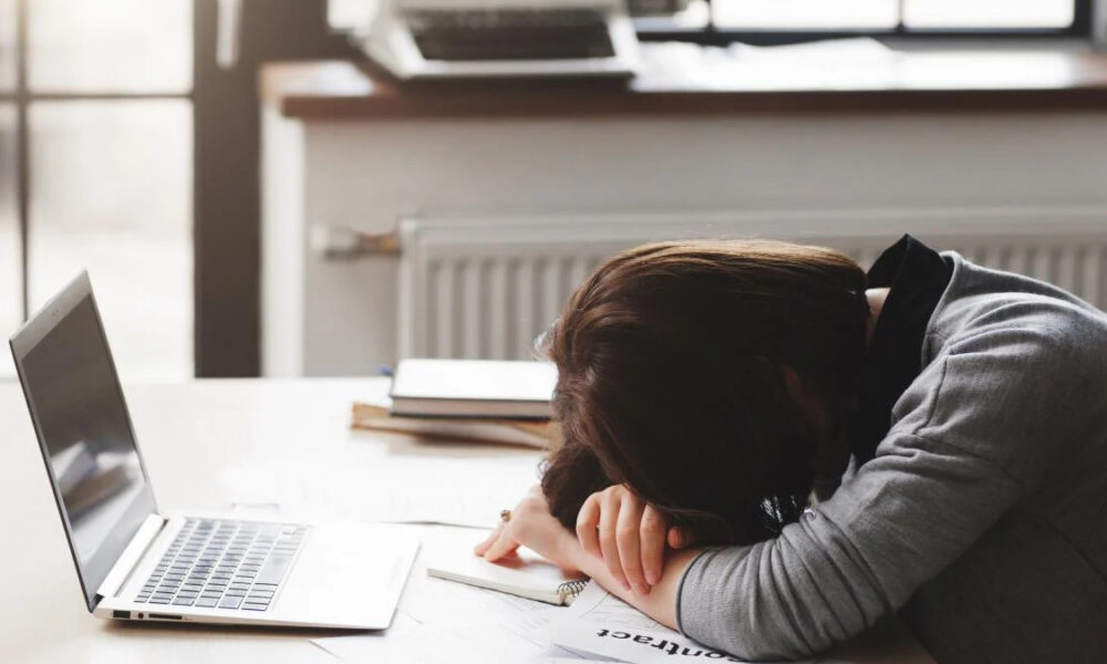 Person rests head on folded arms on a desk beside an open laptop, looking exhausted while studying or working from home.