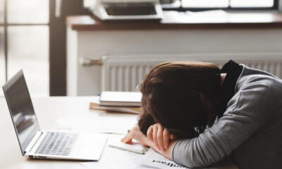 Person rests head on folded arms on a desk beside an open laptop, looking exhausted while studying or working from home.