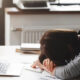 Person rests head on folded arms on a desk beside an open laptop, looking exhausted while studying or working from home.