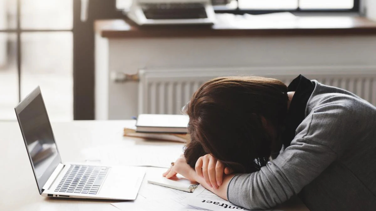 Person rests head on folded arms on a desk beside an open laptop, looking exhausted while studying or working from home.