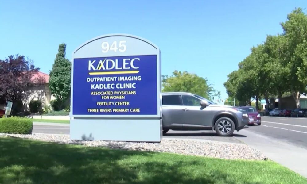 Large blue Kadlec clinic sign with 945 address, surrounded by greenery; a silver SUV is parked to the right on a sunny day.