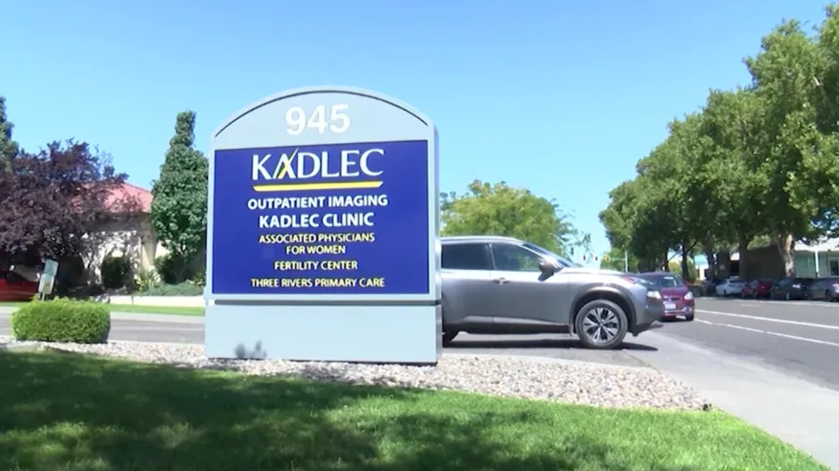 Large blue Kadlec clinic sign with 945 address, surrounded by greenery; a silver SUV is parked to the right on a sunny day.