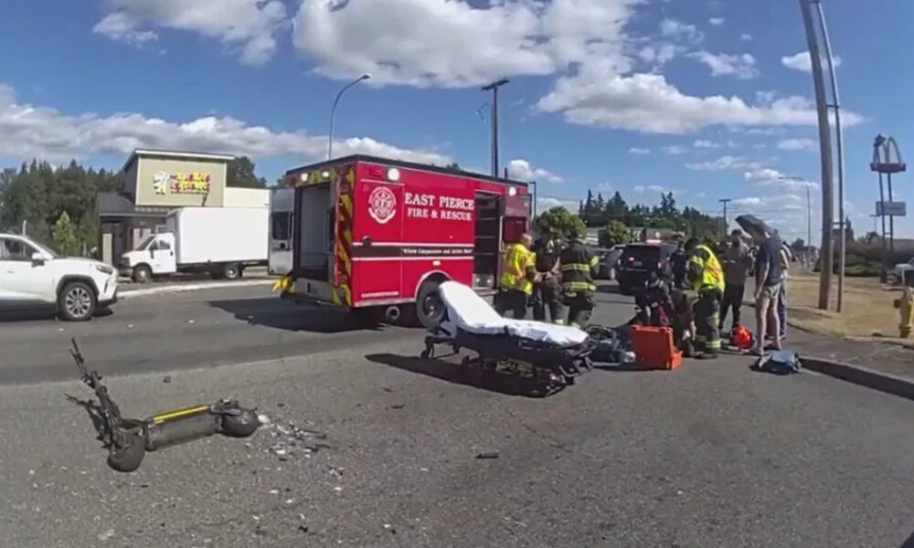 Emergency responders around a red Fire & Rescue ambulance at a multi-vehicle crash, with a gurney and a fallen scooter on the road nearby
