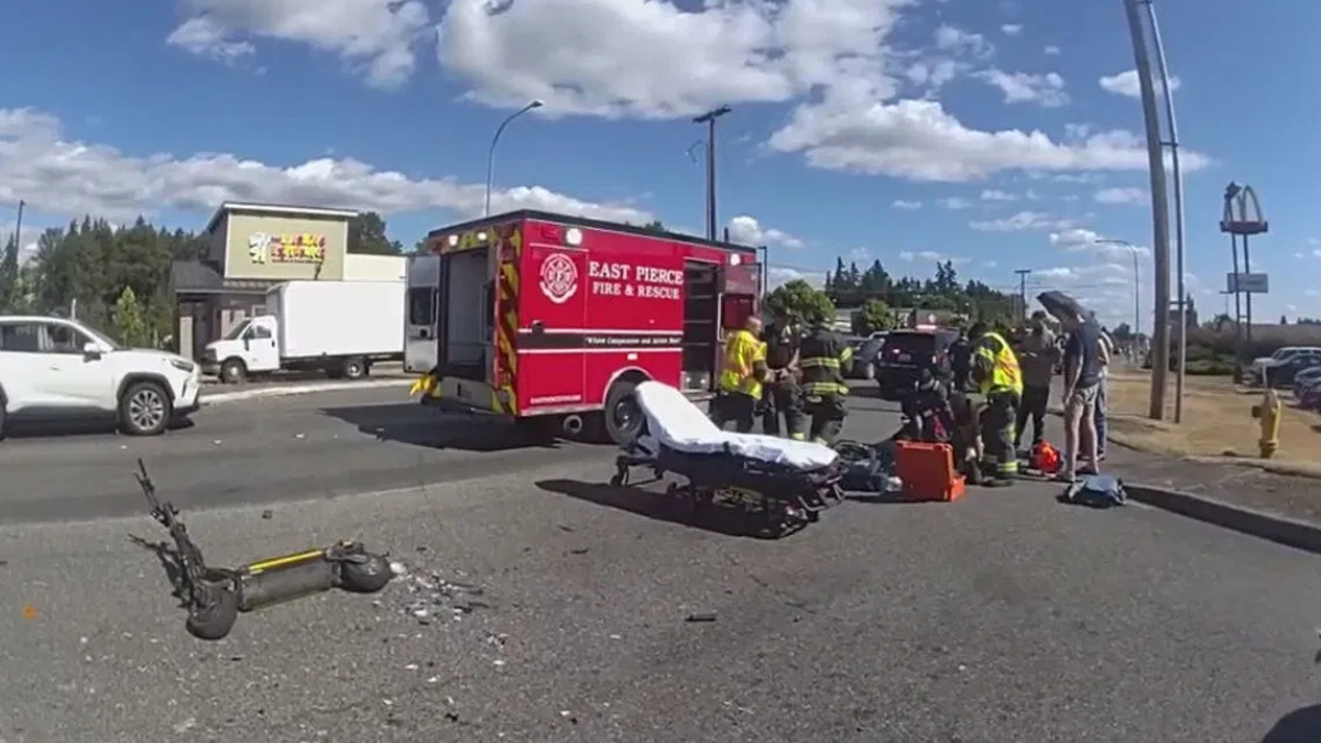 Emergency responders around a red Fire & Rescue ambulance at a multi-vehicle crash, with a gurney and a fallen scooter on the road nearby