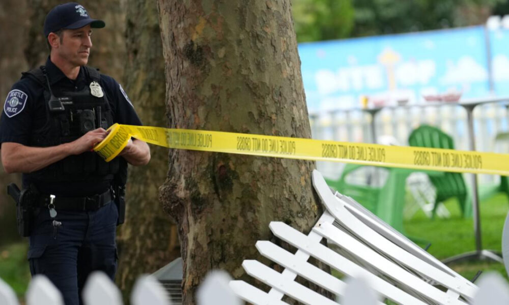 Police officer standing behind yellow caution tape at a park, securing a crime-scene area near a tree and fence pieces ahead of blue banners in the background.